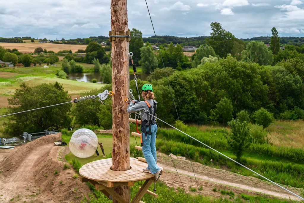 Person iført sikkerhedsudstyr og hjelm forbereder sig til zipline fra en træplatform i Fyns Sommerland, en udendørs oplevelsespark omgivet af grønne områder.